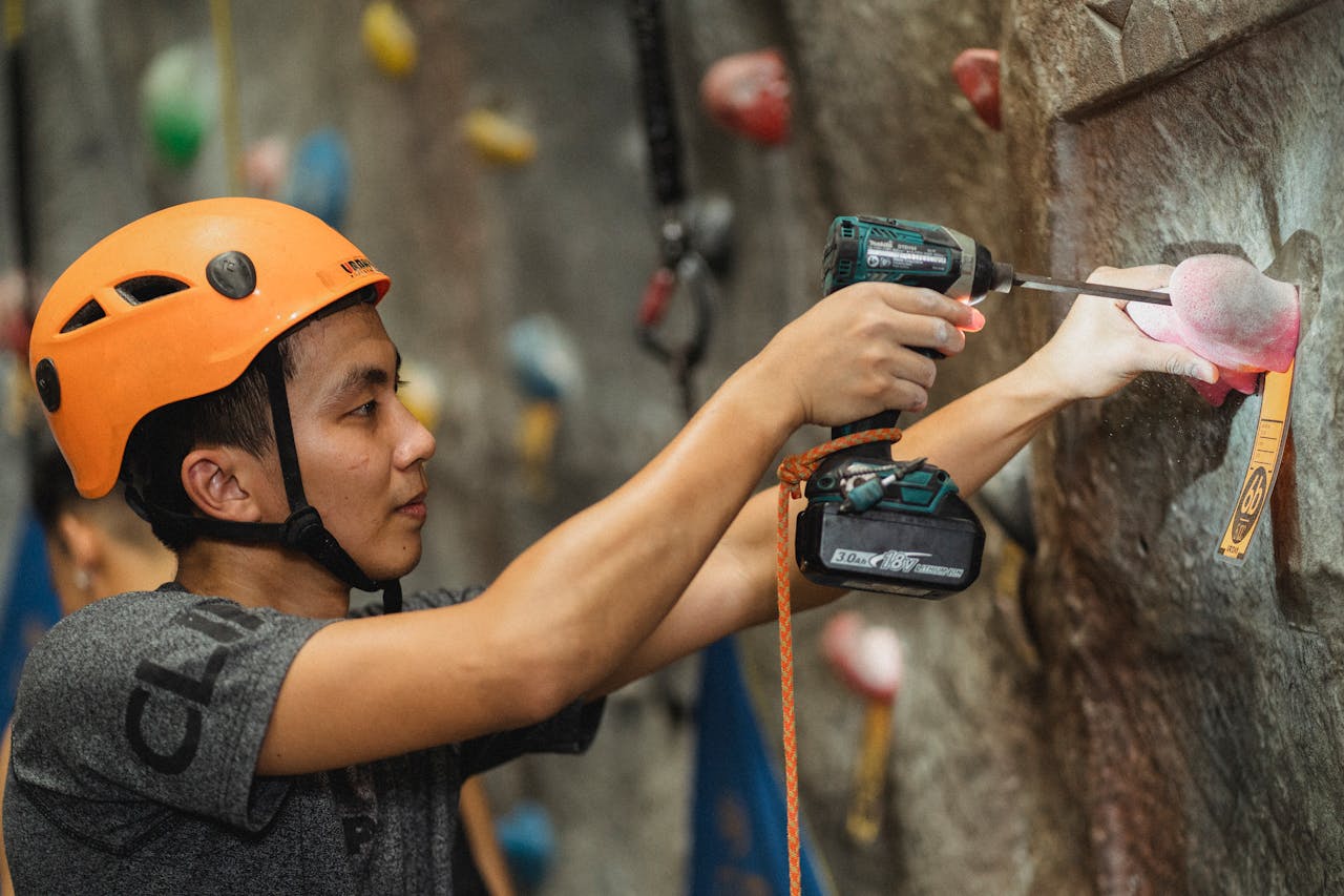 Side view of young workman in orange helmet attaching climbing hold with drill using screwdriver in bouldering gym